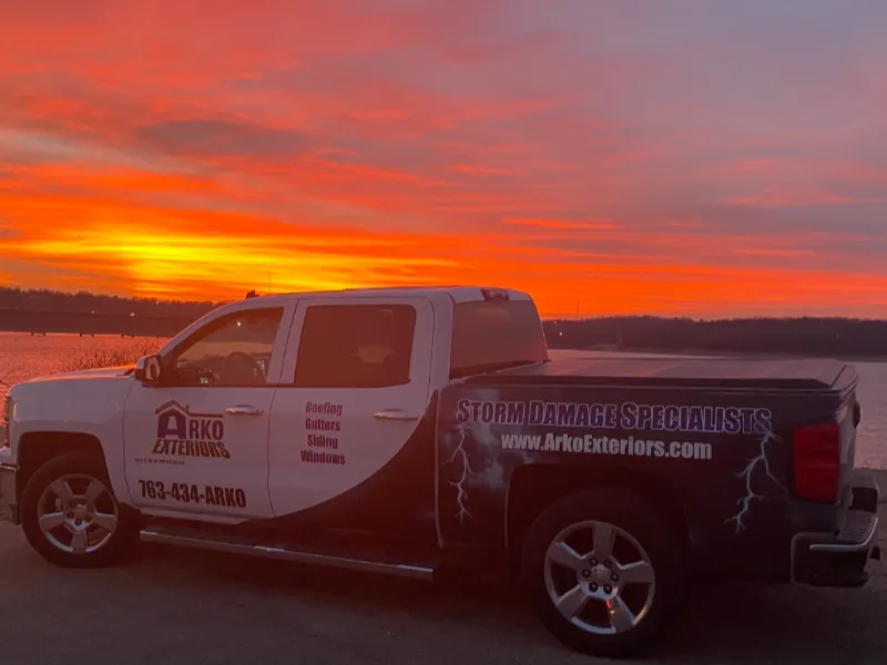 Pickup truck with Arko Exteriors storm damage specialist advertisement parked by a lake at sunset.
