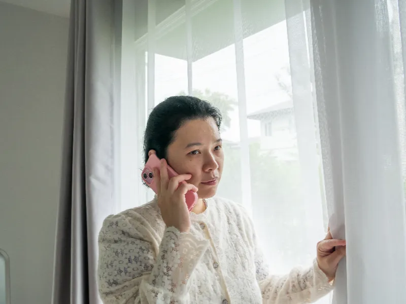A woman in a white lace top talks on a pink phone while looking out a window.