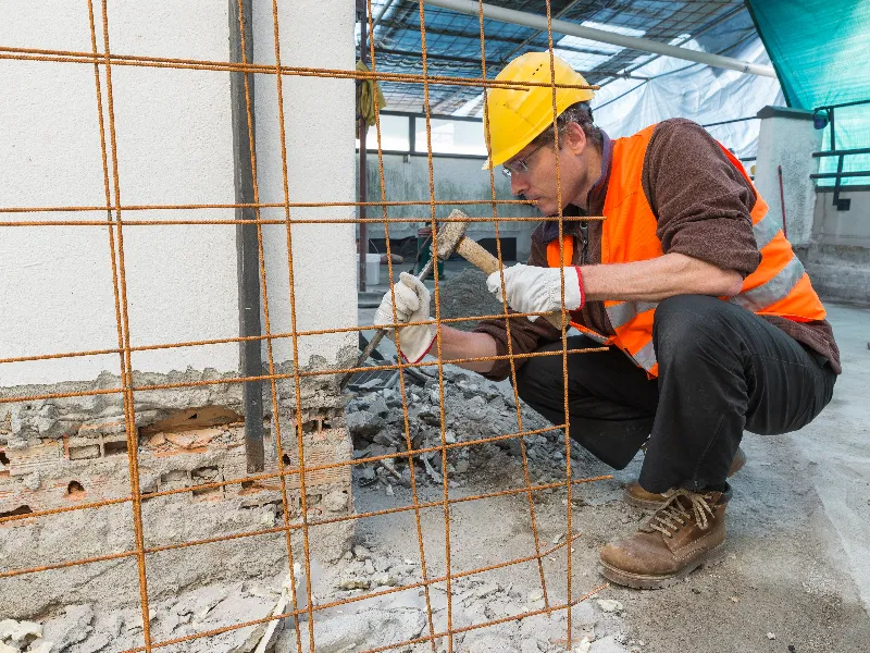 Construction worker in a yellow helmet and orange vest chiseling a wall behind a metal grid.