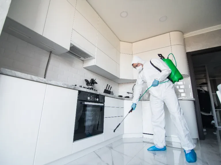 Person in protective suit disinfects a modern white kitchen floor with a sprayer.