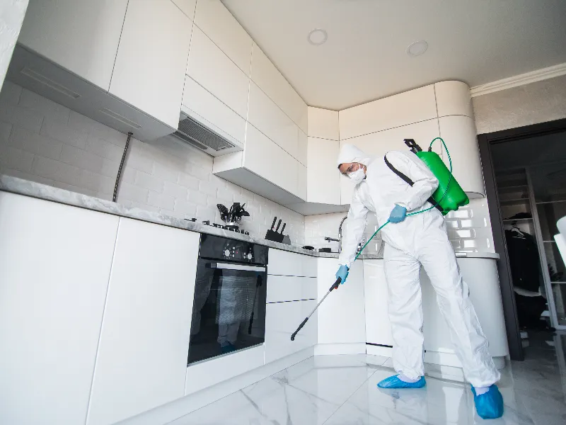 Person in protective suit disinfects a modern white kitchen floor with a sprayer.