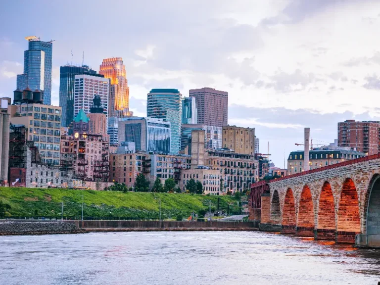 City skyline with modern and historic buildings beside a river and an arched stone bridge at sunset.