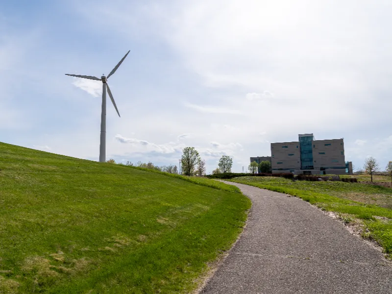 Path leading up a grassy hill toward a wind turbine and a modern building under a cloudy sky.