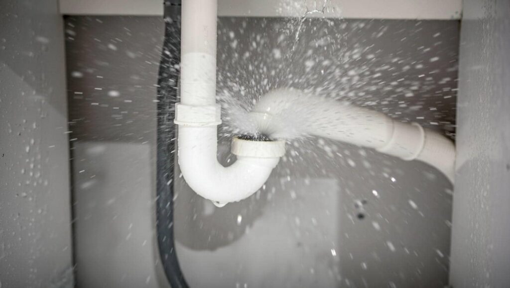White PVC pipe under a sink spraying water from a leak at a joint.