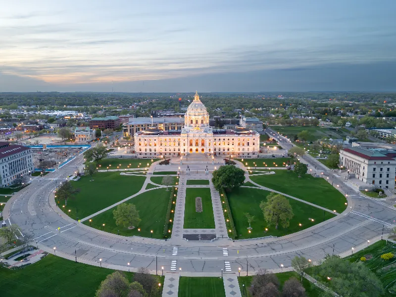 State capitol building illuminated at dusk with surrounding roads and green lawns.