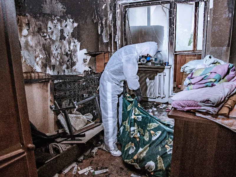 Person in protective suit and mask cleaning debris in a fire-damaged room.