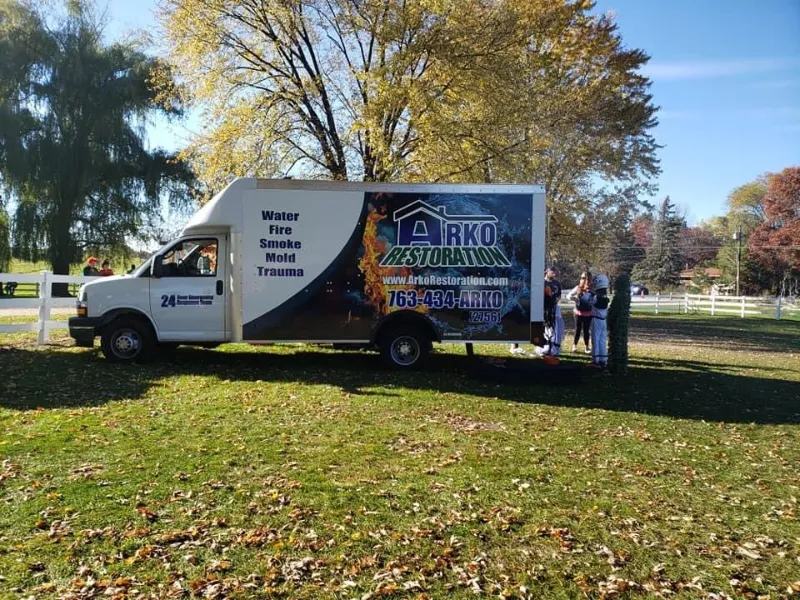 Restoration service truck parked on grass with people standing nearby under autumn trees.