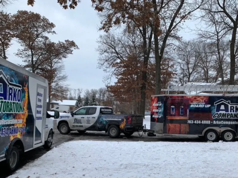 Pickup truck towing a trailer and a box truck with ARKO Restoration branding in a snowy yard.