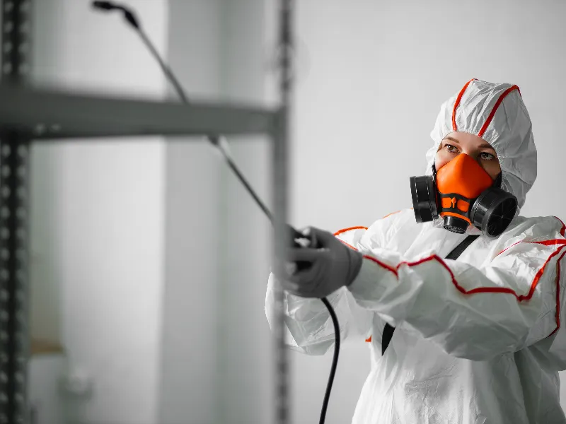 Person in protective suit and orange respirator spraying disinfectant indoors.